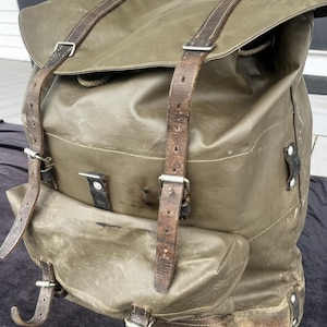May include: An olive green rucksack featuring a large main compartment with a top flap, a smaller front pocket, and brown leather straps with metal buckles. The bag has a weathered appearance with visible wear on the leather and fabric surfaces.
