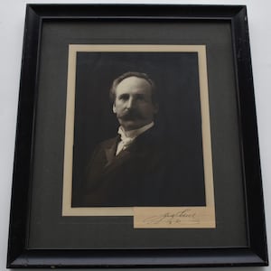 May include: A framed black and white portrait of a man with a large mustache, wearing a dark suit and a white shirt with a bow tie. The portrait is in a black frame with a cream-colored matting.