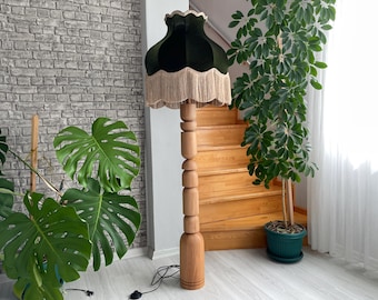 May include: A wooden floor lamp with a dark green velvet lampshade and beige fringe. The lamp has a stacked, cylindrical wooden base. A large green plant is to the right of the lamp.