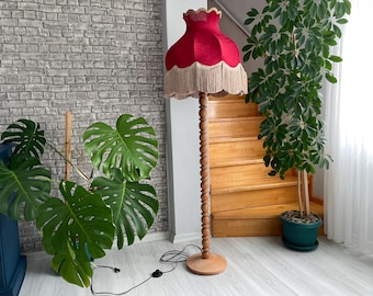 May include: A vintage-style floor lamp with a red lampshade and a wooden base. The lampshade has a scalloped edge and fringe. The lamp stands approximately 60 inches tall. The lamp is in a room with plants and a wooden staircase.