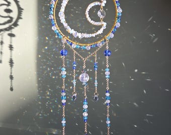 May include: A circular suncatcher featuring a gold-toned wire frame with a crescent moon shape inside. The design is adorned with blue, clear, and iridescent beads, with five dangling chains of matching beads and a clear crystal point at the bottom.