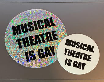 May include: Two round buttons with the text "Musical Theatre is Gay" printed in black on a white background. One button is covered in iridescent glitter.