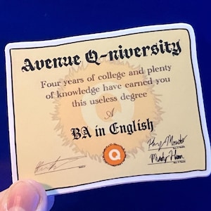 Puede incluir: Adhesivo de título universitario de Avenue Q. El adhesivo es una Licenciatura en Inglés. El texto dice: "Cuatro años de universidad y mucha sabiduría te han dado este título inútil". El adhesivo está sostenido por una mano.