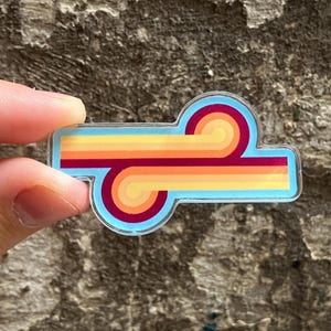 May include: A clear acrylic pin featuring a horizontal design with two parallel, curved lines in shades of yellow, orange, and deep red. The graphic is set against a light blue background, creating a smooth, rounded shape with a glossy finish.