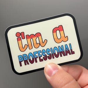 May include: A rectangular sticker with rounded corners and a cream-coloured background. The text "i'm a" appears in a gradient of red, orange, and yellow, while the word "PROFESSIONAL" is written in blue and dark red, all enclosed in a black border.