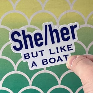 Könnte beinhalten: Ein weißer Aufkleber mit dem Text "She/her BUT LIKE A BOAT" in Marineblau. Der Aufkleber wird von einer Hand vor einem Hintergrund mit einem grün-gelben Schuppenmuster gehalten.