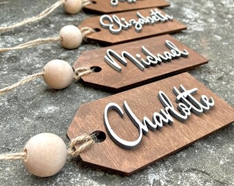 Könnte beinhalten: Vier Holz-Namensschilder mit den Namen Jessica, Elizabeth, Michael und Charlotte in weißer Schrift. Jedes Etikett ist braun mit einer natürlichen Holzperle und einer Schnur aus Bindfaden befestigt. Die Etiketten sind auf einer strukturierten grauen Oberfläche angeordnet.