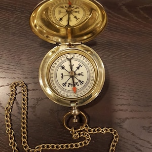 May include: A gold compass with a chain attached. The compass has a round face with black markings and a red needle. The compass is open and the lid is flipped over.
