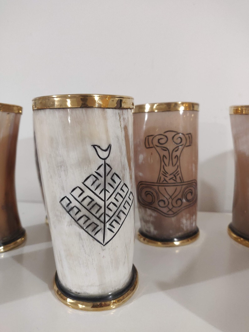 May include: Set of four drinking horns with gold rims and black etched designs. The designs include a tree, a hammer, and a geometric pattern.