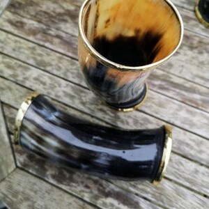 May include: Two drinking horns with gold trim. One horn is upright and the other is lying down. Both horns are made of dark brown and black material.