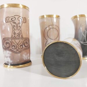 May include: Four drinking horns with gold rims and different etched designs. The horns are made of a light brown material and are sitting on a white surface.