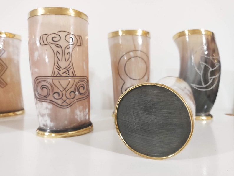 May include: Four drinking horns with gold rims and different etched designs. The horns are made of a light brown material and are sitting on a white surface.
