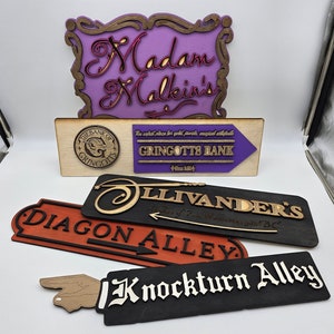 Puede incluir: Cinco letreros de madera con varios nombres de tiendas con temática de Harry Potter. El letrero superior es morado con las palabras "Madam Malkin's" en una fuente cursiva. El siguiente letrero es marrón con las palabras "The Bank of Gringotts" y un pequeño logotipo circular con las palabras "The Bank of Gringotts". El tercer letrero es negro con las palabras "Ollivanders" y un gráfico de varita. El cuarto letrero es rojo con las palabras "Diagon Alley". El quinto letrero es negro con las palabras "Knockturn Alley".