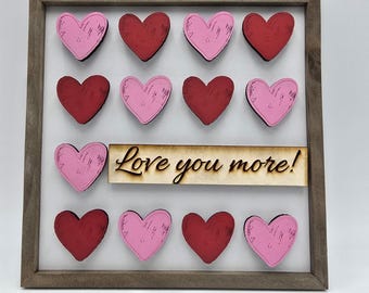 May include: A framed wooden sign with a white background and 12 wooden hearts in alternating red and pink colors. The sign says "Love you more!" in a wood-burned font.
