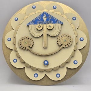 Puede incluir: Una pieza decorativa de madera con un diseño de cara sonriente. La cara tiene un techo azul y dorado, ojos circulares y una nariz vertical. La pieza circular tiene un borde festoneado y detalles en azul y dorado. La combinación de colores es dorada, crema y azul. Un artículo artesanal encantador.