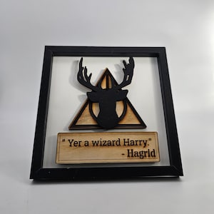 Puede incluir: Una pieza de arte de pared de madera con marco negro con una silueta negra de una cabeza de ciervo dentro de un triángulo con un círculo en el centro. La cita "Yer a wizard Harry." - Hagrid está escrita en una placa de madera debajo del ciervo.