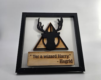 May include: A black framed wood wall art piece with a black silhouette of a stag's head inside a triangle with a circle in the center. The quote "Yer a wizard Harry." - Hagrid is written on a wooden plaque below the stag.