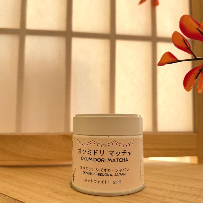 May include: A cylindrical white metal tin of Okumidori Matcha tea with a light pink label. The label features black text reading Okumidori Matcha, Origin: Shizuoka, Japan, and Net Weight: 30g, along with decorative scalloped line details.