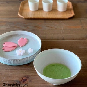 May include: A white ceramic bowl containing frothy green matcha tea, a light blue plate with three pink petal-shaped sweets and two small white flower-shaped candies, and three white cups on a wooden tray. The text Akimatcha.com is visible.