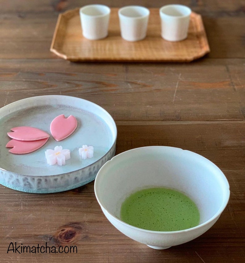 May include: A white ceramic bowl containing frothy green matcha tea, a light blue plate with three pink petal-shaped sweets and two small white flower-shaped candies, and three white cups on a wooden tray. The text Akimatcha.com is visible.