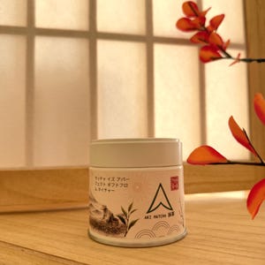May include: A cylindrical white metal tin of Aki Matcha tea featuring a label with a line drawing of a person harvesting tea leaves, a sun icon, and a green triangle logo. The text reads "Aki Matcha" and includes Japanese characters.