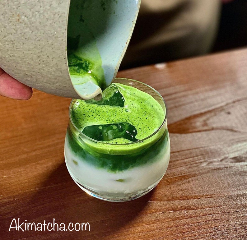 May include: A glass of iced matcha latte with a layer of green matcha foam being poured from a ceramic jug. The drink is on a wooden surface.