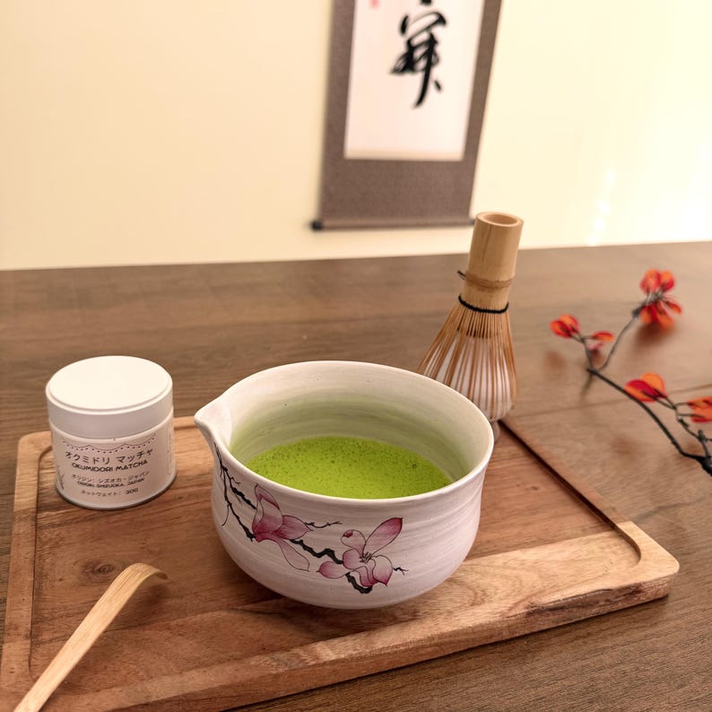 May include: A white ceramic matcha bowl with a pink floral design containing bright green tea, a white cylindrical tin labeled Okumidori Matcha, a bamboo whisk, and a curved bamboo scoop arranged on a rectangular wooden serving tray.