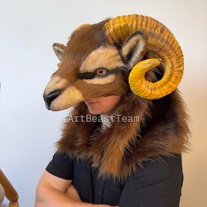 May include: A detailed ram mask with large, curved, yellow-gold horns. The mask features brown and tan fur, with the text "ArtBeastTeam" visible. The mask is worn over a black shirt.