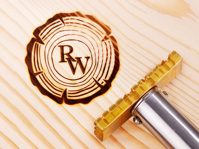 May include: A close-up of a wood burning tool with a brass head and a silver handle. The tool is used to create a wood-burned design on a wooden surface. The design is a circle with the letters "R" and "W" inside.