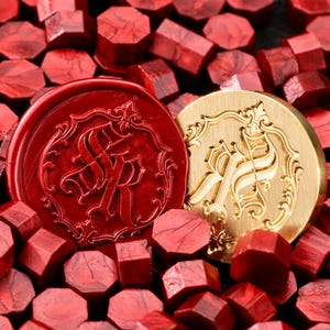 May include: A circular brass wax seal stamp featuring an ornate monogram of the letters FR surrounded by a decorative border, positioned next to a matching red wax seal impression, all resting amidst a pile of small, hexagonal red wax beads.