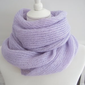 Puede incluir: Una bufanda de punto de color lila claro hecha de un hilo suave y delicado. La bufanda está tejida de forma holgada, creando una sensación aireada y ligera.