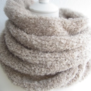 Op de afbeelding: Een beige infinity-sjaal met een getextureerde, krullende en geluste gebreide constructie. De sjaal is in meerdere lagen gewikkeld, waardoor een volumineuze, cirkelvormige vorm ontstaat die rond een witte cilindrische presentatievorm ligt.