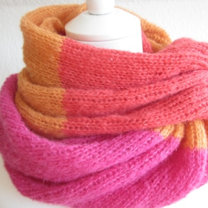 Op de afbeelding: Een roze, oranje en gele gebreide sjaal. De sjaal is gevouwen en gedrapeerd over een wit mannequin.