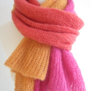 May include: A long, hand-knitted scarf featuring a loose, open-stitch texture. The scarf is color-blocked with vibrant sections of bright orange, coral pink, and deep magenta, draped in multiple layers around a white display form.