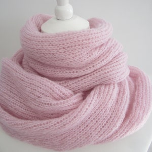 Puede incluir: Una bufanda infinita de color rosa claro hecha de hilo tejido suave, aireado y semitransparente. La bufanda presenta una textura de punto abierto y holgado, drapeada en múltiples capas circulares que crean un aspecto voluminoso y fruncido.