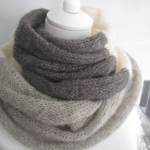 Peut inclure: Une écharpe infinie en tricot épais présentant un design color-block dans des tons crème, beige clair et gris anthracite foncé. L'écharpe est confectionnée dans un fil doux et duveteux avec une texture tricotée lâche créant un aspect volumineux.