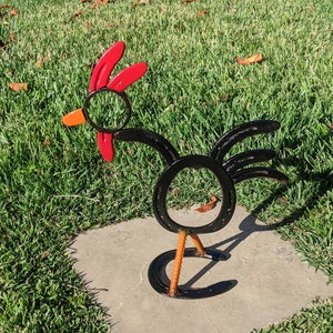 May include: A black metal rooster sculpture made from horseshoes. The rooster has a red comb and wattle, orange legs, and a black body. The sculpture is standing on a gray concrete slab.