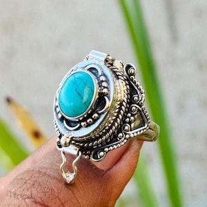 May include: A silver ring with an oval turquoise stone. The ring has an ornate design with rope and bead detailing around the stone. The ring is held up by a finger against a blurred green and white background.