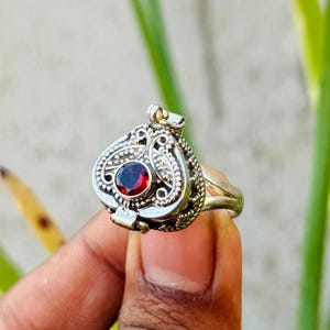 Peut inclure: Bague en argent avec un pendentif orné en forme de cœur. Le pendentif présente une petite pierre précieuse rouge et ronde au centre, entourée de détails de filigrane complexes. La bague est tenue dans une main sur un fond vert et gris flou.