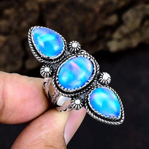 May include: A silver ring featuring three oval and teardrop-shaped opals. The opals have a vibrant blue color with iridescent qualities. The ring has a detailed, ornate design with a textured border around each stone.