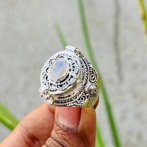 May include: Ornate silver ring featuring a central oval-shaped moonstone. The ring has detailed filigree and scrollwork around the stone and band. The ring is held up against a blurred green and gray background.