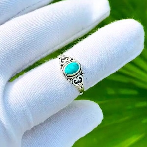 May include: A silver ring featuring an oval-shaped turquoise stone set in a decorative metal band with intricate scrollwork and beaded detailing. The ring is displayed on a finger, showcasing the polished metal finish and the vibrant blue-green color of the stone.