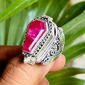 May include: A silver ring with a large, rectangular, bright red gemstone. The ring has intricate, detailed patterns around the gemstone and on the band. The ring is held up against a background of green leaves.