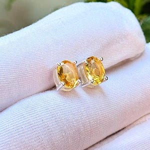 Könnte beinhalten: Ohrstecker mit ovalen, goldgelben Edelsteinen in Silberfassung. Die Ohrringe werden auf einer weißen Oberfläche präsentiert, mit grünen Blättern im Hintergrund. Die Edelsteine haben eine warme, strahlende Farbe.