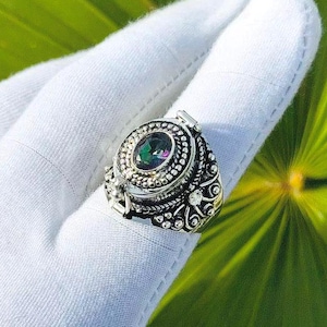 May include: A silver ring with an oval, iridescent gemstone. The ring has an ornate design with small, clear stones around the main stone. The ring is displayed on a white gloved hand against a green leaf background.