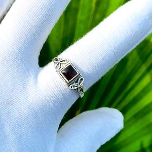May include: A silver ring featuring a square-cut dark red gemstone set in a decorative metal band with intricate filigree detailing on the sides. The ring is displayed on a finger covered by a white glove, showcasing the polished metal finish.