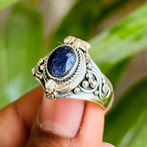 Könnte beinhalten: Ein silberner Ring mit einem ovalen, dunkelblauen Edelstein. Der Ring hat aufwendige Filigranarbeiten um den Stein und auf dem Band. Der Ring wird in einer Hand vor einem verschwommenen grünen Hintergrund gehalten.