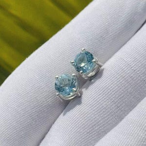 May include: A pair of light blue gemstone stud earrings. The earrings have a round cut and are set in a silver metal. The earrings are displayed on a white surface with a green background.