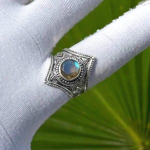 Pode incluir: Um anel de prata com uma pedra preciosa de labradorite redonda e facetada com iridescência azul. A banda de metal tem um padrão em relevo intrincado com detalhes de contas que rodeiam a pedra numa forma de escudo com bordas pontiagudas.