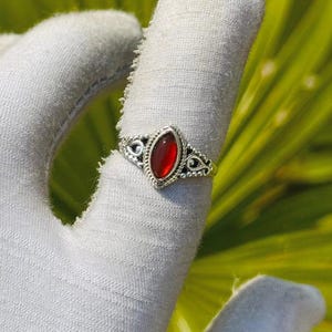 May include: A silver ring with an oval, deep red gemstone. The ring has an ornate, filigree design around the stone and on the band. The ring is displayed on a white surface against a backdrop of green foliage.